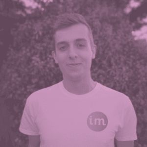 Portrait of Joe Lamyman, smiling with his brown hair swept to the side. The photo captures Joe
from the torso up. Joe is wearing a white t-shirt with a rounded logo, feature the letters 'im' on the left breast.
Joe is stood in front of a large bush, covering the background. A pink and purple filter has been applied to the
photo.