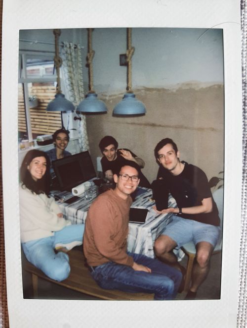 Jade site behind a computer monitor at the top of the table smiling. To the right of Jade is Matt, holding a dice up to the camera. To the right of Matt, at the bottom of the table, Joe holds a pitcher full of cocktails. To the left of Joe sits James. Between James and Jade, Siobhan sits smiling at the camera. On the table is a computer monitor with a map barely visible. In the background, a newly plastered wall provides a grungy backdrop to the photo.