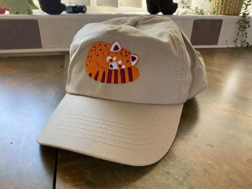 A beige cap sitting upon our stone coloured kitchen countertop. On the front of the cap is an embroidered red panda. The red panda is tucked up into a ball, it’s large bushy tail wrapped around the front of it’s body, with it’s head nestled into it’s tail. The panda’s eyes are closed in a way that show delight, with them arched. The red panda has dark brown stripes on the tail and a few dark brown markings on it’s light orange body.