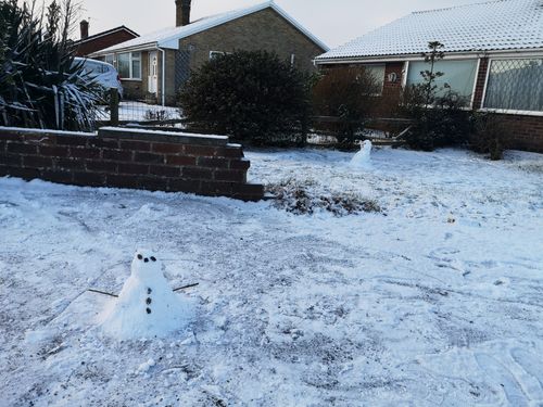 A small snowperson (roughly 20cm tall) sits at the front of the image with two stick arms, coal eyes, a coal nose and three coal buttons. The ground is covered in a thin blanket of snow. In the background is the snowperson we built previously. The rest of the background is made up by a small brick wall surrounding some grass, a series of bushes and our bungalow.